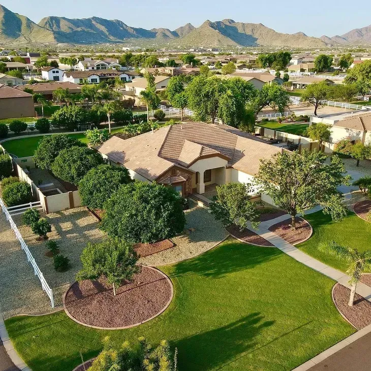 Aerial view of a suburban home with a green lawn, trees, and desert mountains in the background.