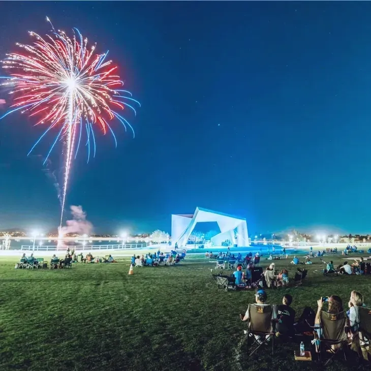 Fireworks over a nighttime outdoor concert crowd gathered on a grassy field near a lit stage