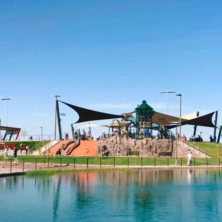 Playground with slides and climbing rocks beside a calm pond under a clear blue sky