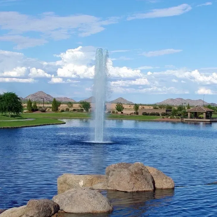 Fountain喷涌 on a blue pond with rocks in front, green lawns, homes, and mountains under a clear sky