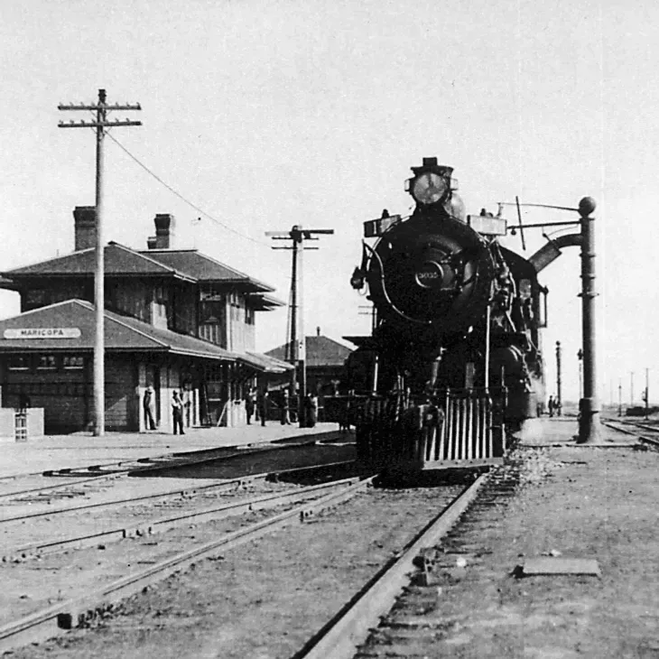 Black-and-white steam locomotive approaching a train station platform with buildings and telegraph poles nearby