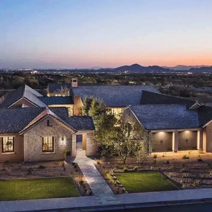 Modern stone house at dusk with lit windows, landscaped yard, and distant mountain skyline