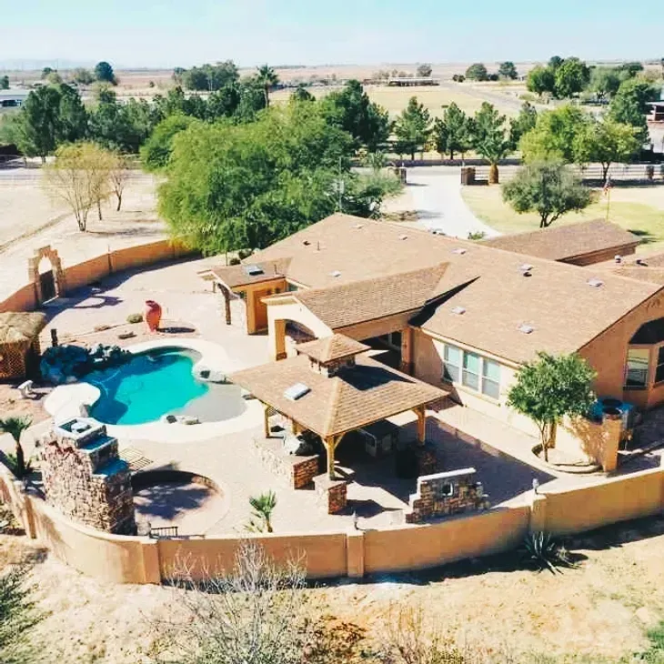 Aerial view of a tan house with a curved backyard pool and patio in a dry rural landscape.