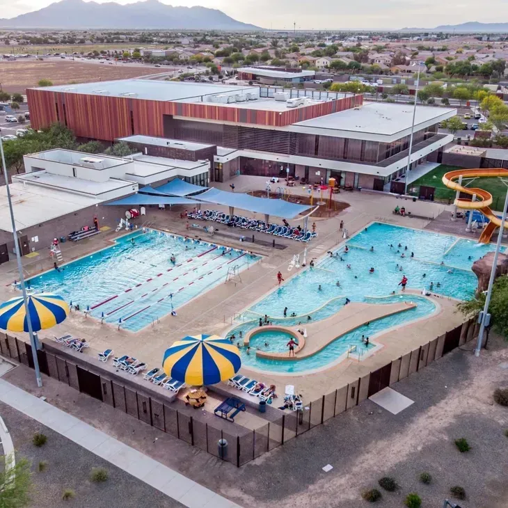 Aerial view of a community pool complex with swimmers, blue water, and a circular splash pool under a striped umbrella.