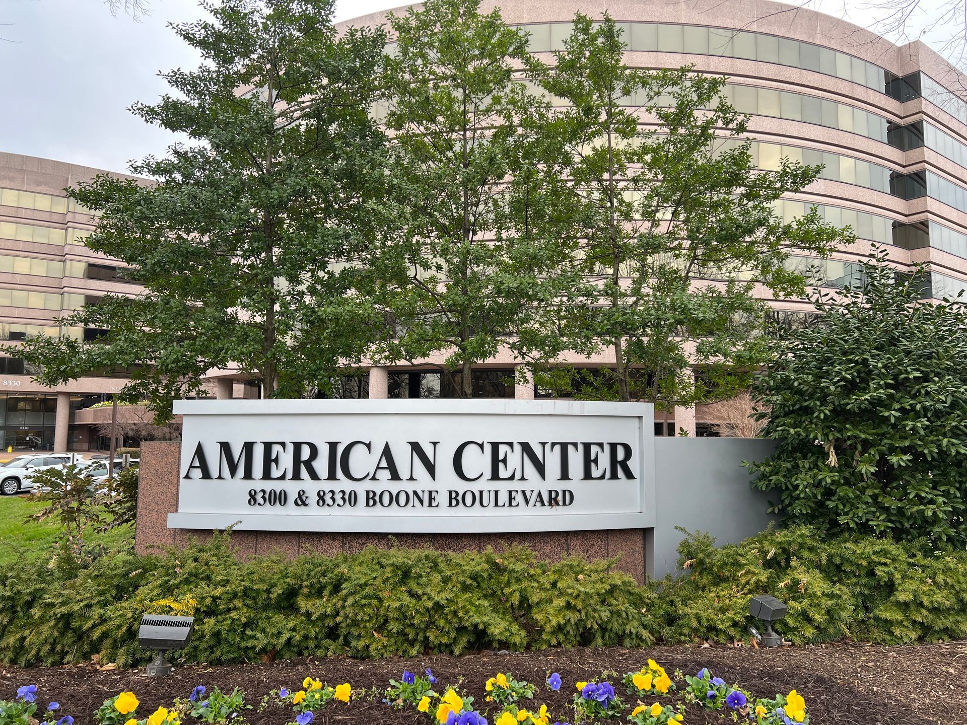 Sign for American Center, 5300 & 5350 Boone Boulevard, with building in the background. Lush greenery and flowers in foreground.