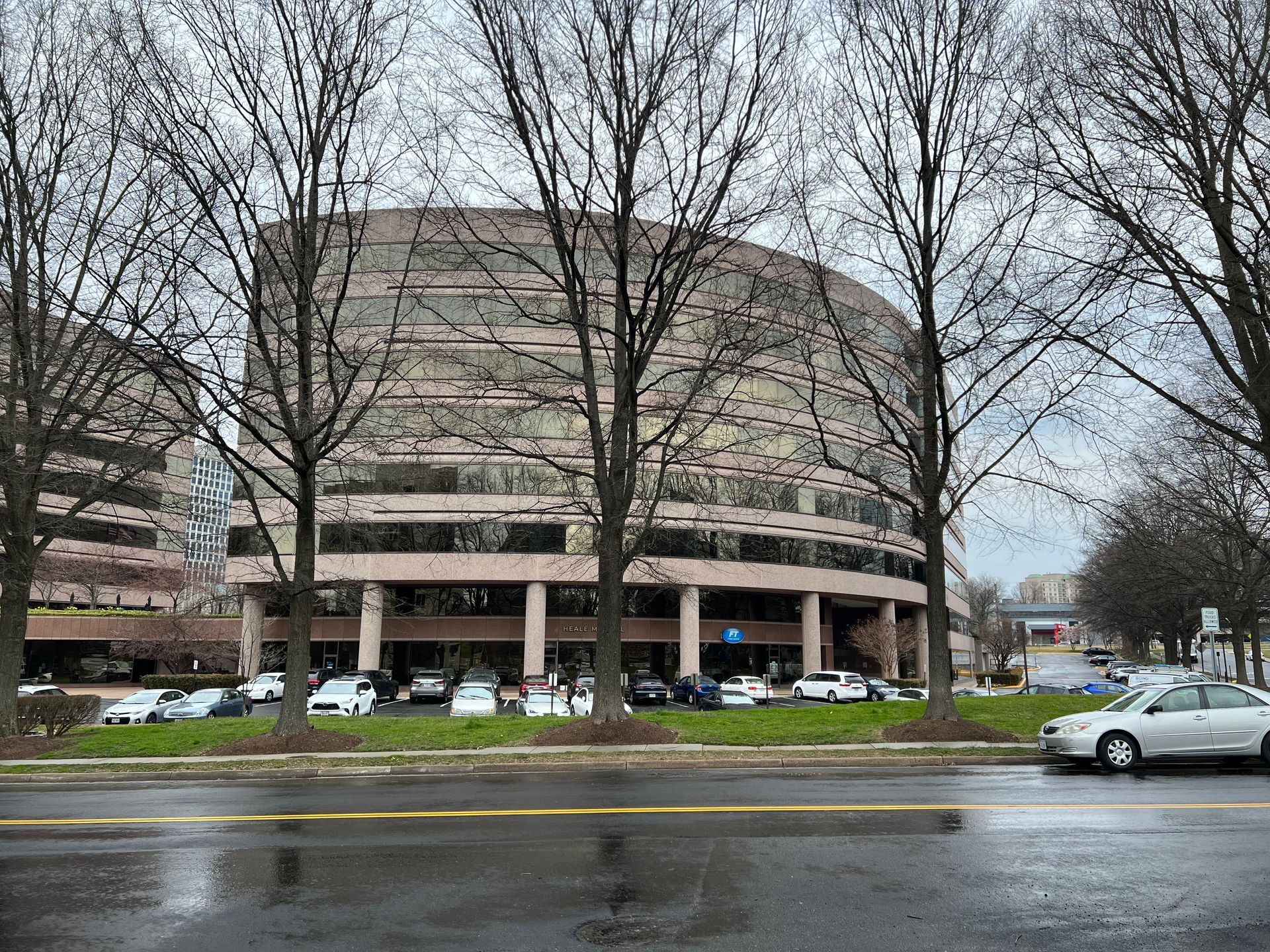 Curved office building behind bare trees and a grassy median, with cars parked in front. Gray sky overhead.
