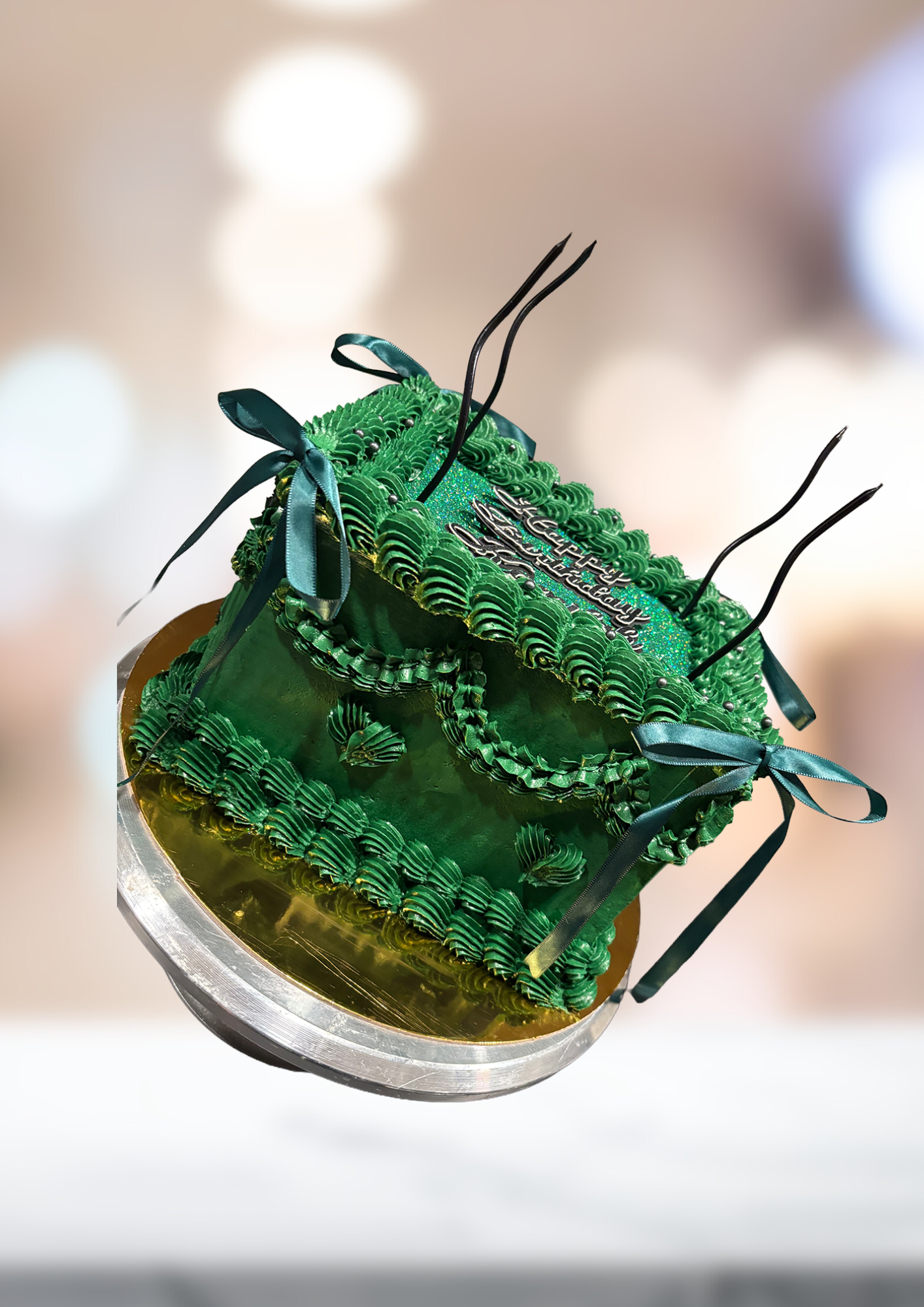 Green frosted cake with black decorative stems on a silver board, against a blurred background