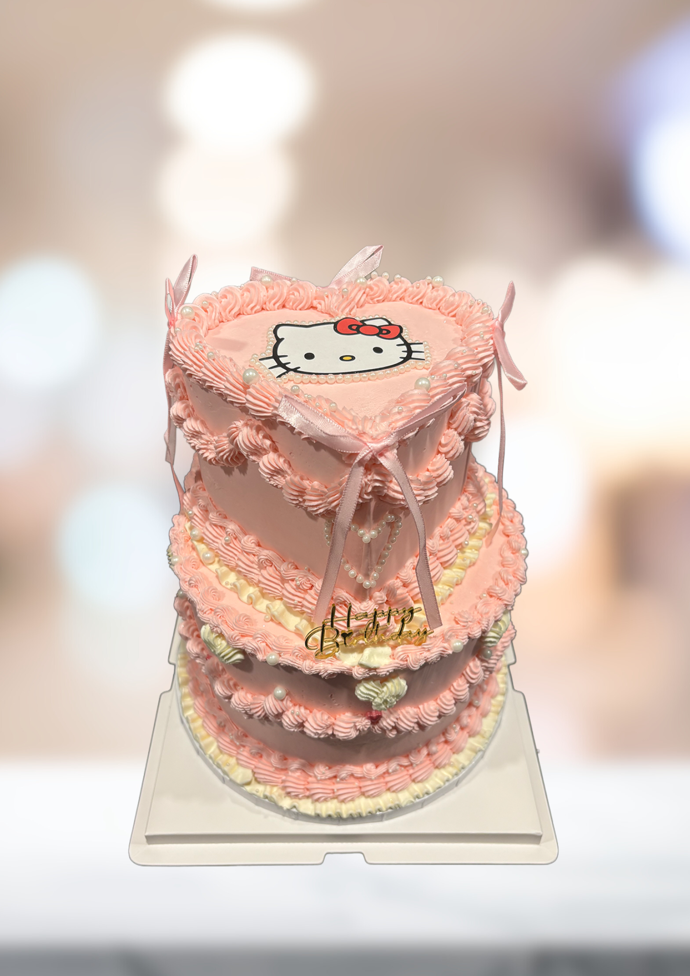 Pink tiered Hello Kitty cake with frilly trim and a bow, on a white stand against a blurred background