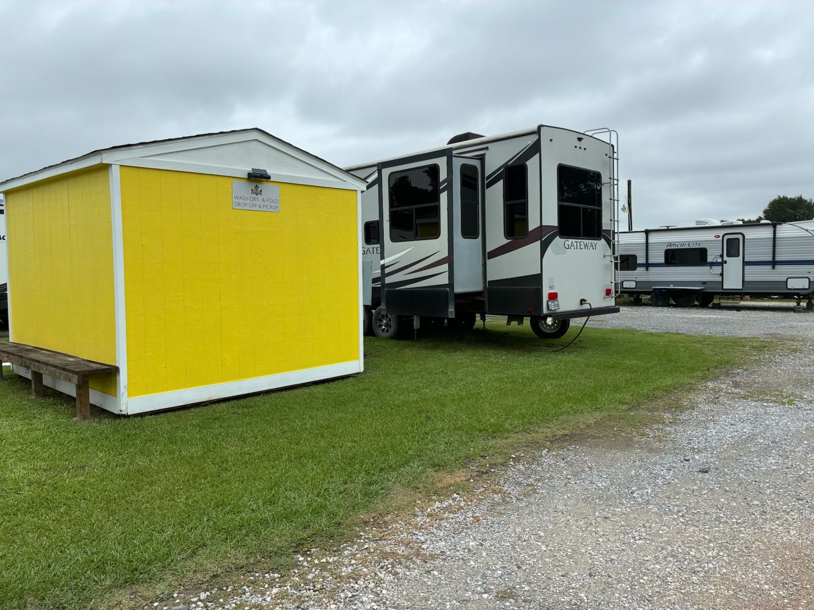 Yellow storage shed beside white RV trailers on a grassy lot with gravel foreground