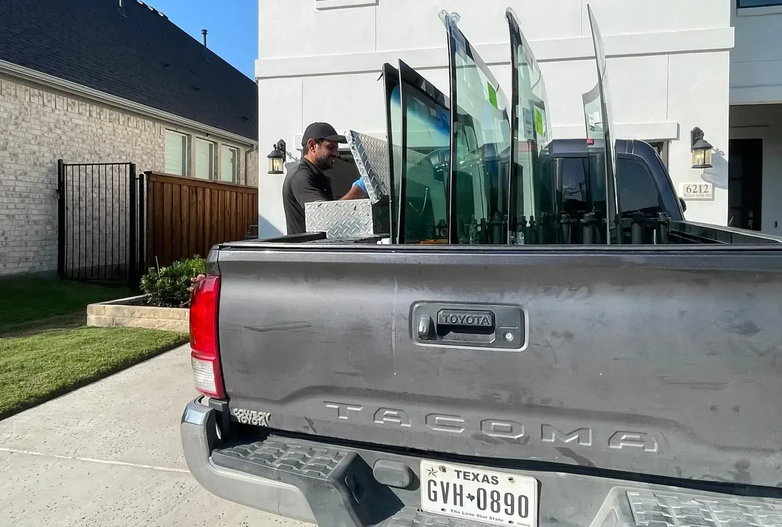 A technician in a black shirt and grey shorts removes a large glass windshield from the bed of a grey pickup truck.