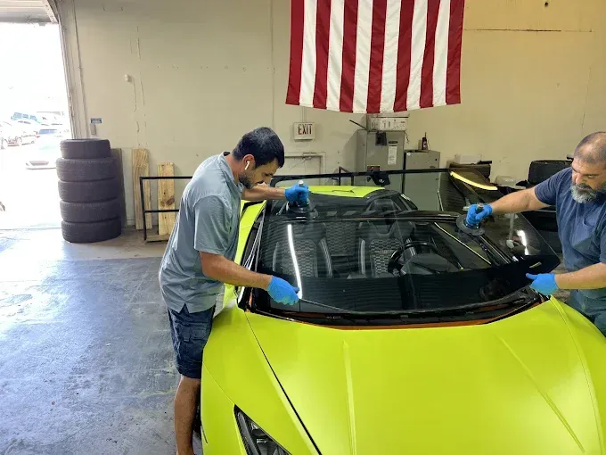 Two people installing a windshield on a lime-green sports car indoors, US flag hanging above.
