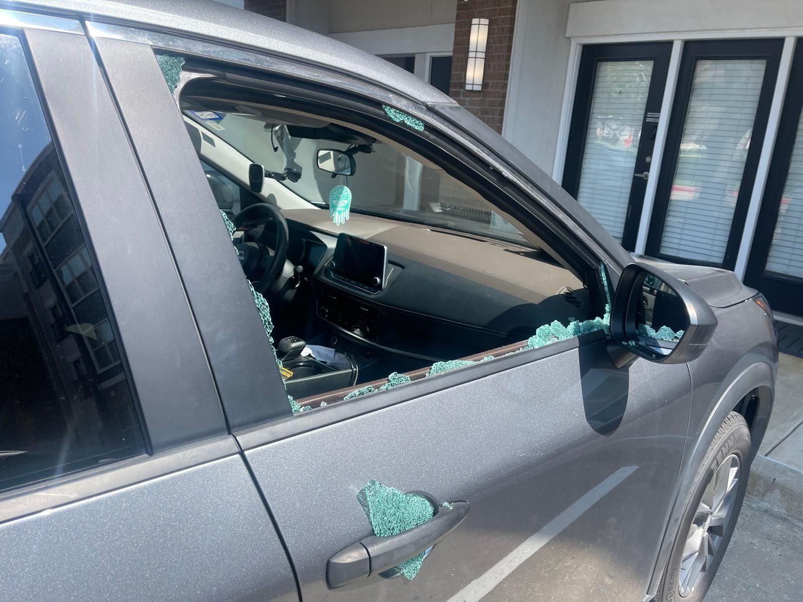 Gray car with shattered driver's side window, parked near a building with black doors.