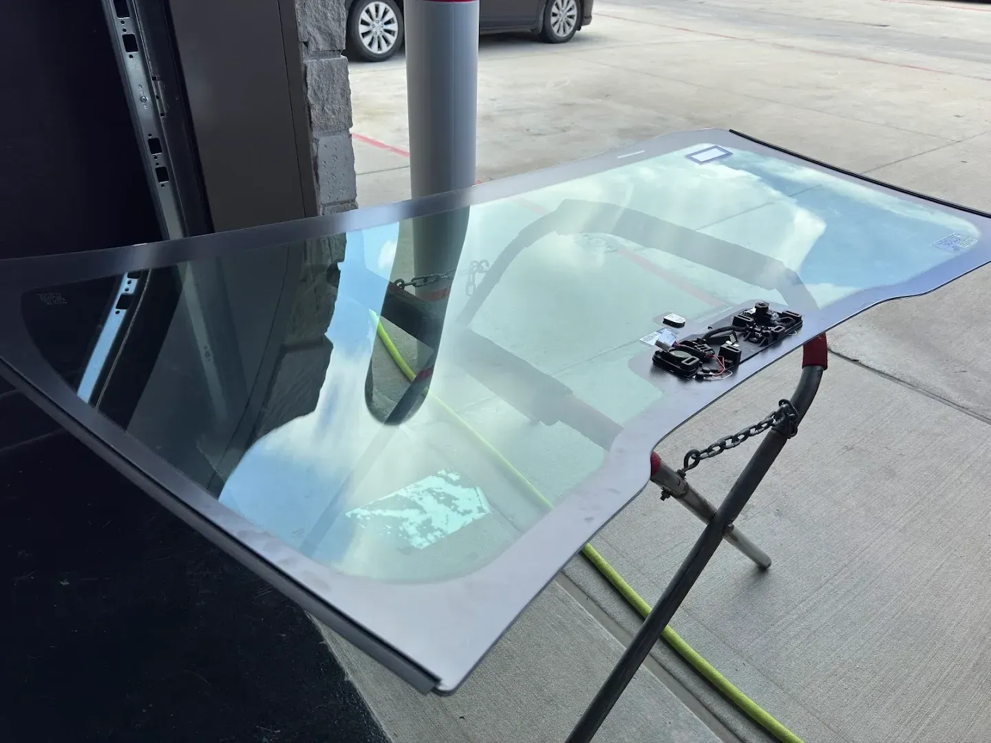Windshield on a stand outside, reflecting the sky and a car. Tools are on top of the glass.