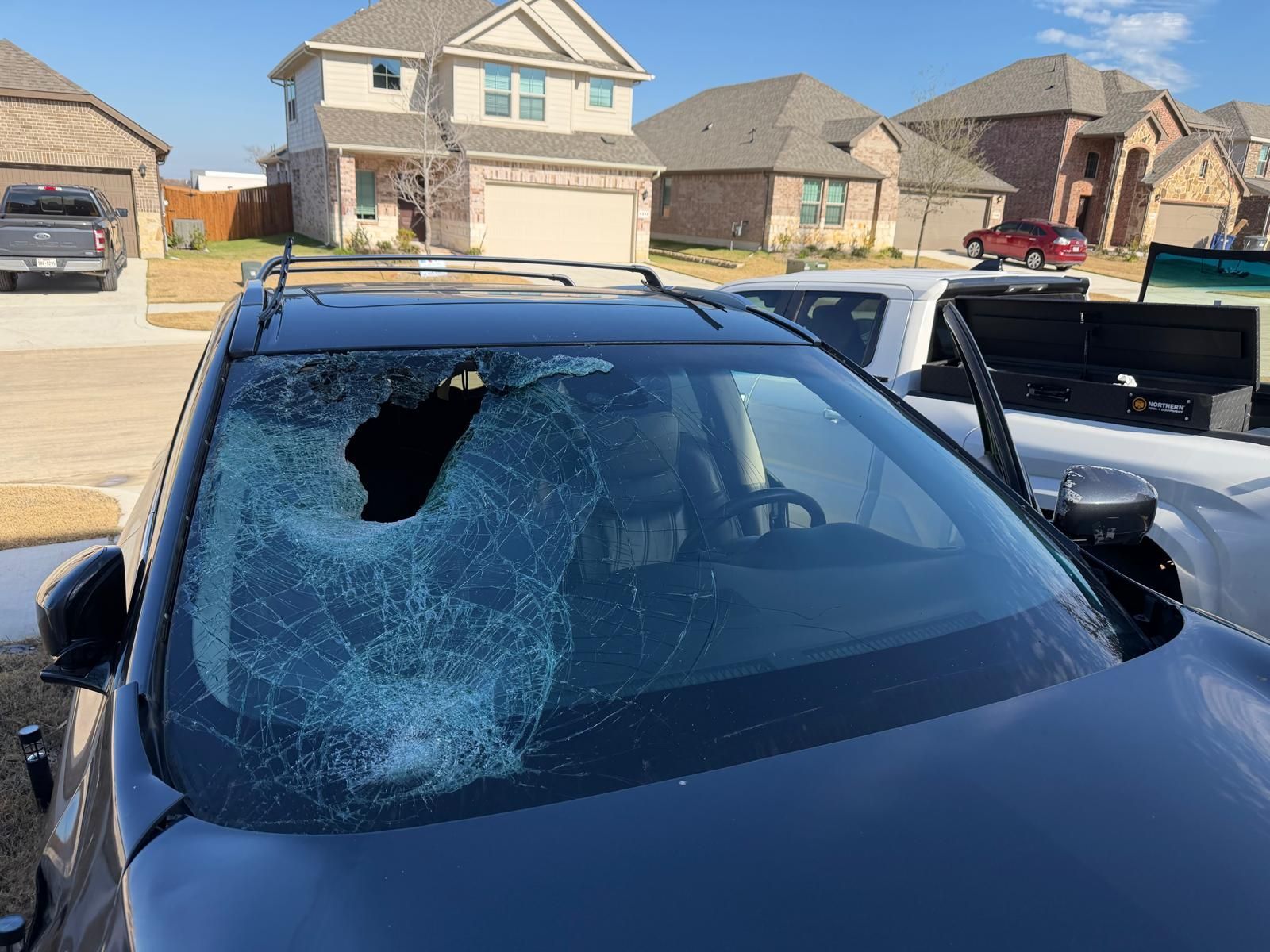Blue car with shattered windshield and large hole, parked in a suburban driveway.