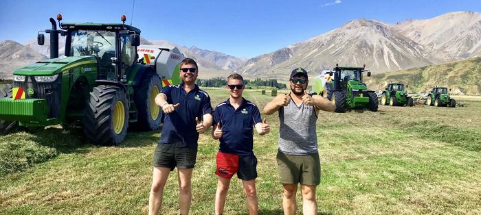 Three men are standing in a field with tractors in the background.