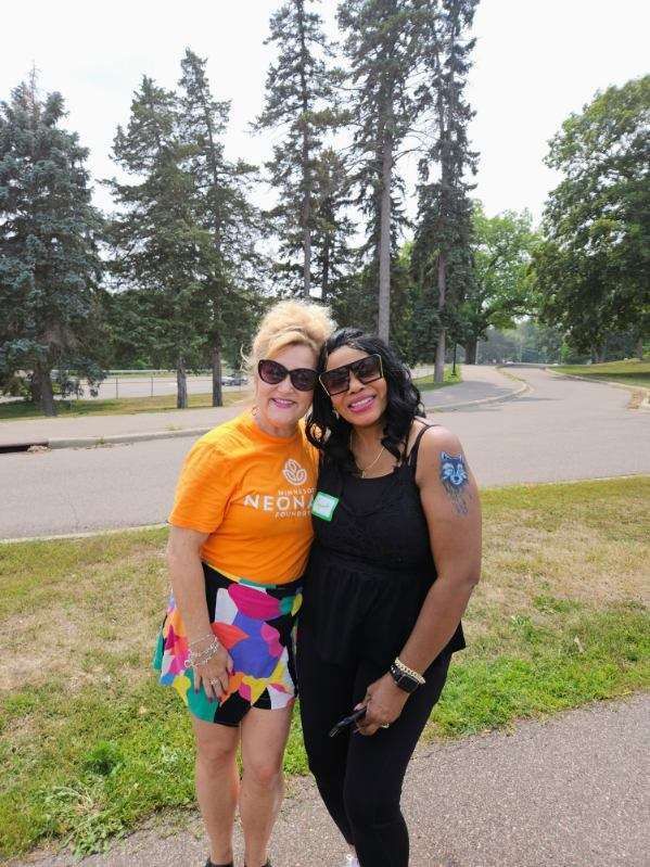 Two women smiling and posing outdoors; one in orange shirt and skirt, the other in black outfit. Trees in the background.