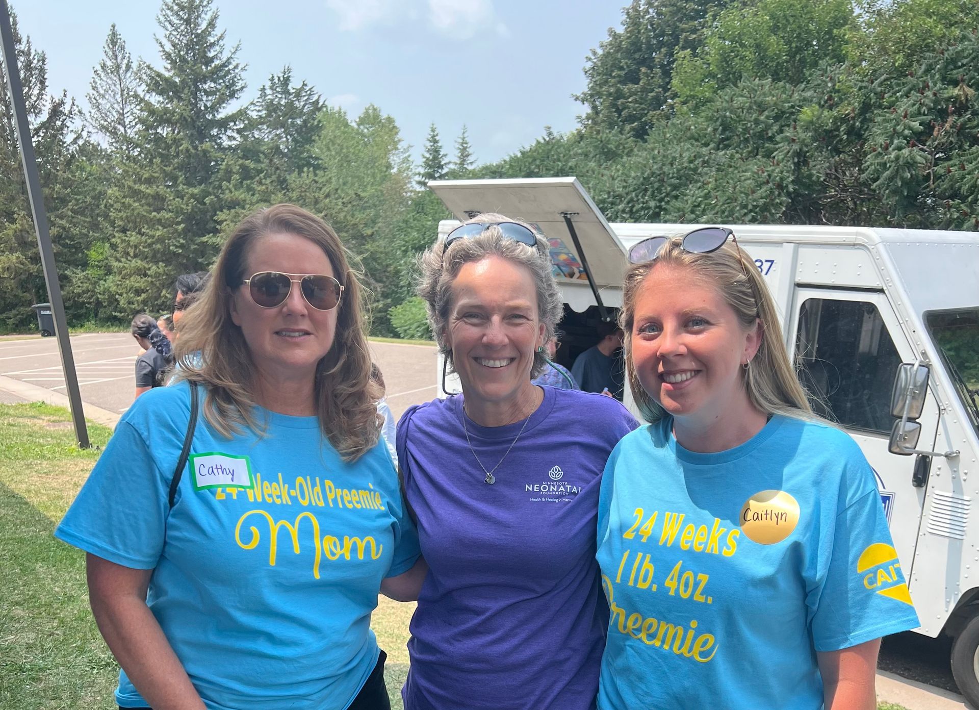 Three women smiling near an ice cream truck. Two wear blue shirts with phrases about a 