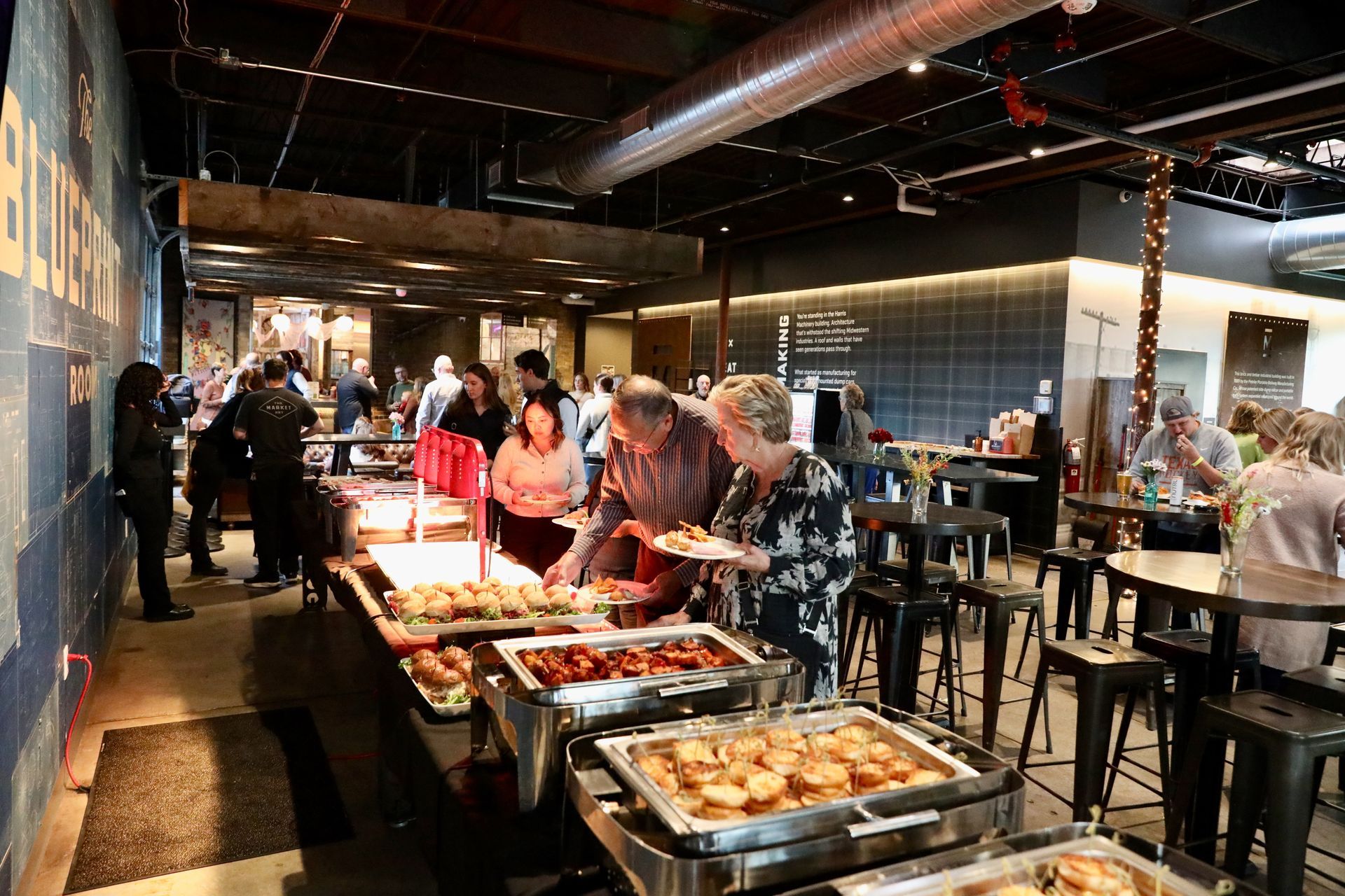 People serving themselves food from a buffet line in a brightly lit indoor venue.