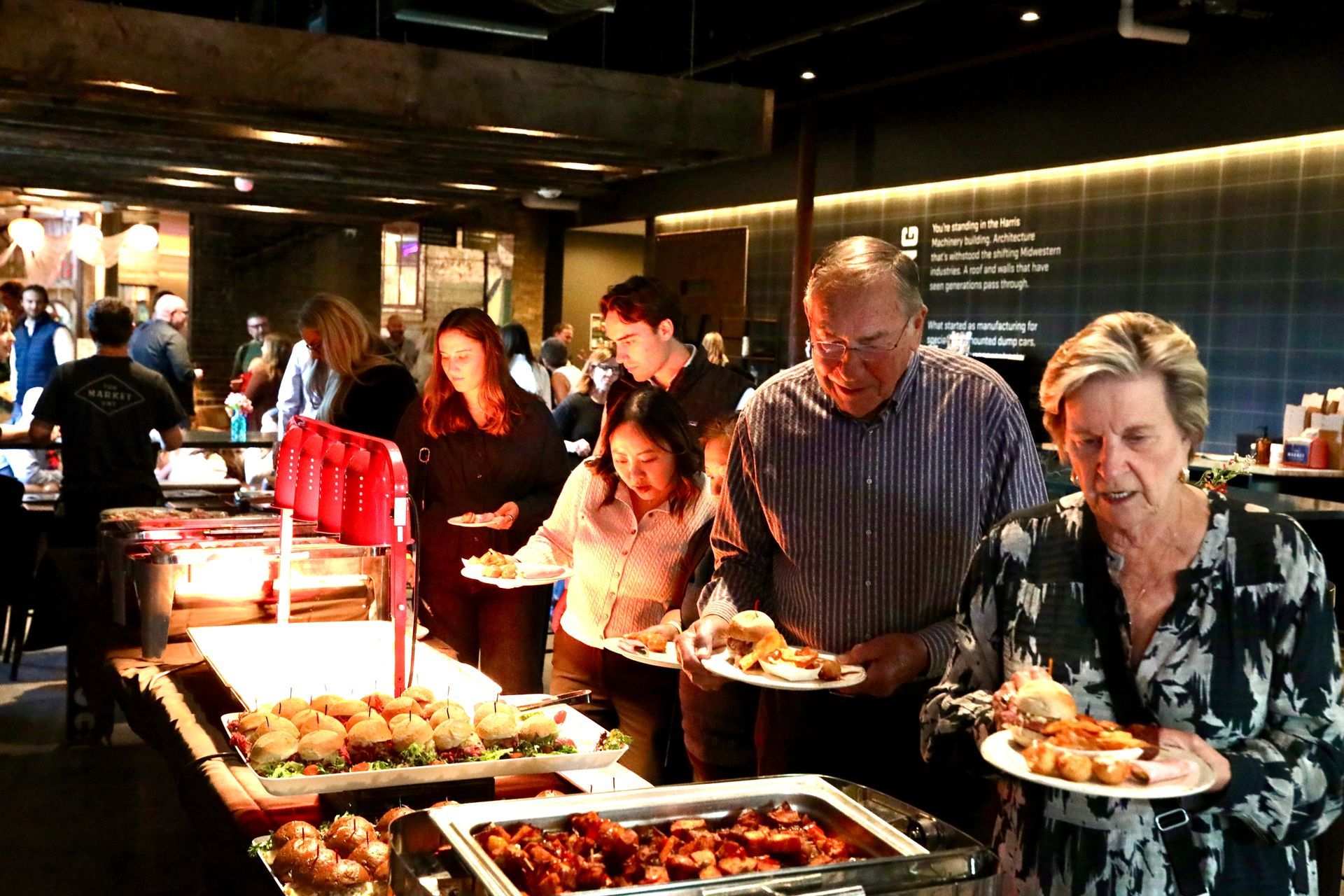 People at a buffet, selecting food. Indoors, warm lighting. Several individuals holding plates.