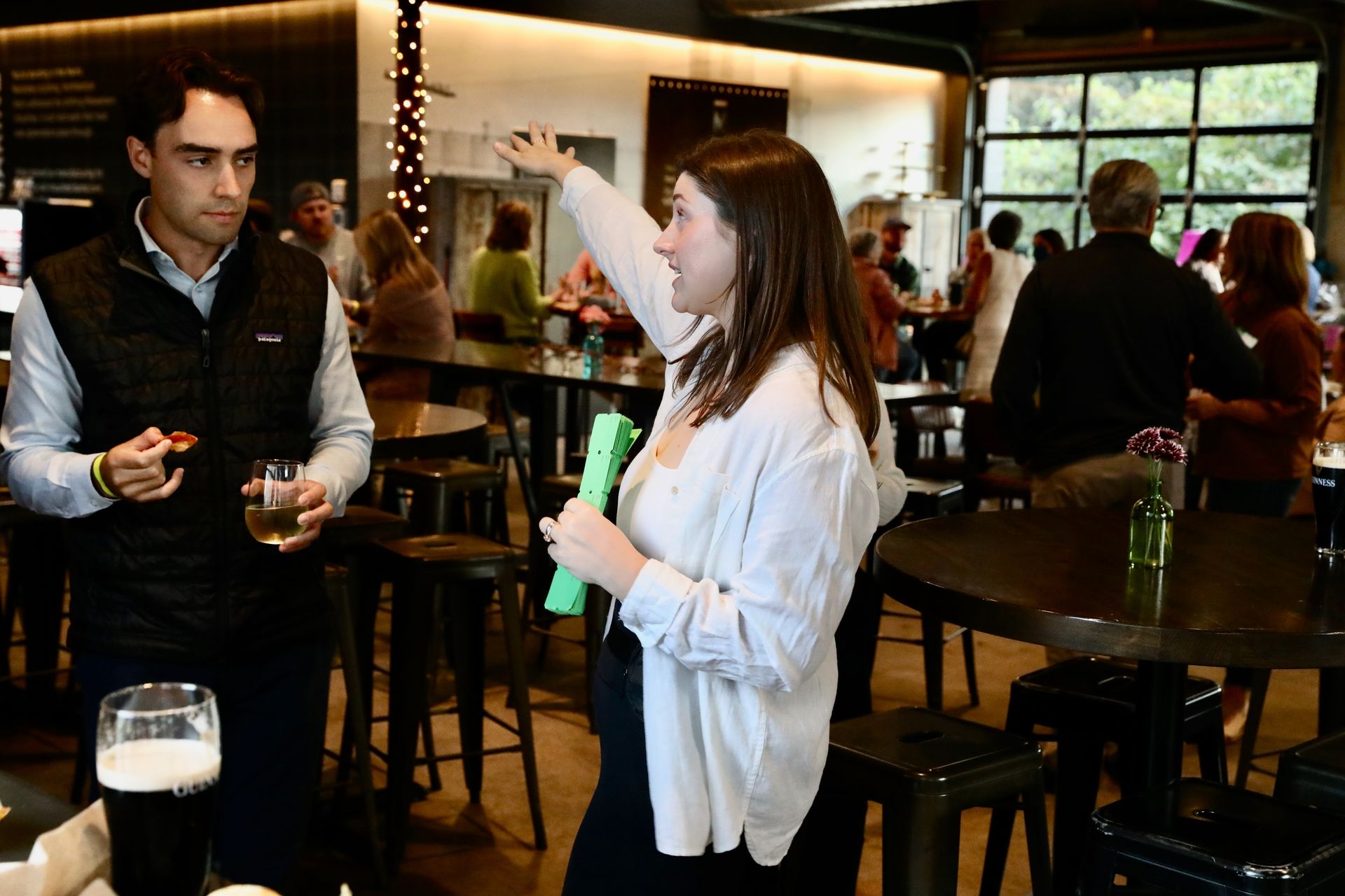 Woman pointing, gesturing, speaking to man, both holding drinks, inside a crowded bar.