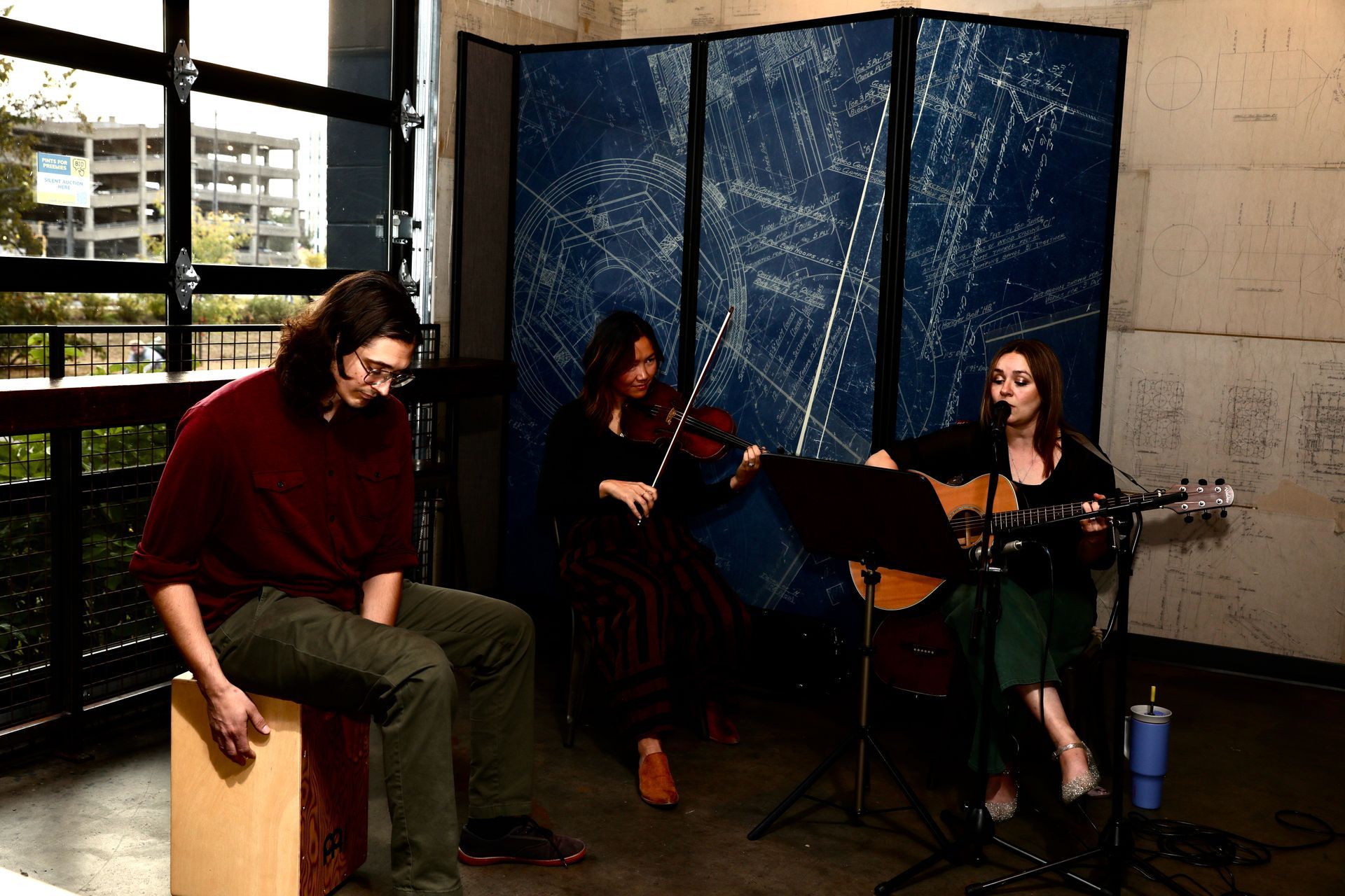 Three musicians performing indoors: a guitarist, a violinist, and a percussionist playing a cajón.