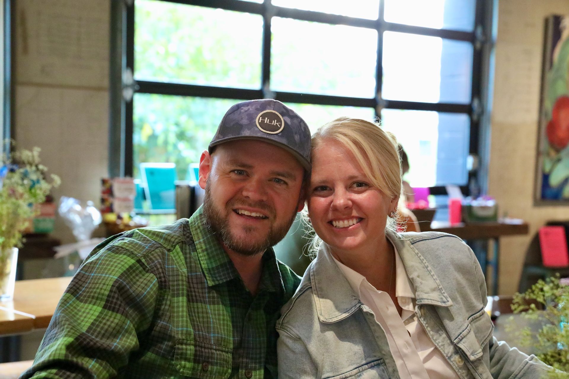 Smiling couple indoors, man in cap and plaid shirt, woman in denim jacket, near a window.