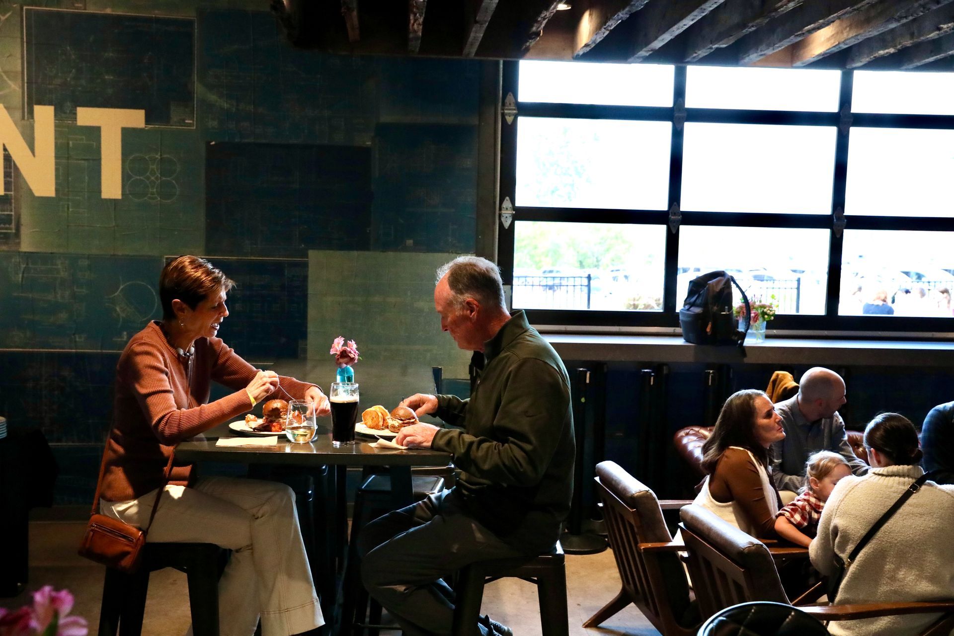 Couple dining at a restaurant, eating and talking. Other patrons in the background.