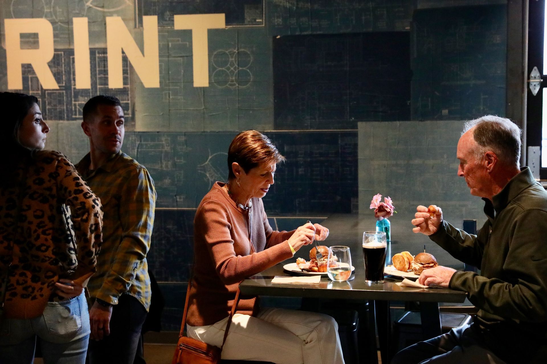 People at a table in a restaurant. Woman taking a photo of a burger; man gestures. 
