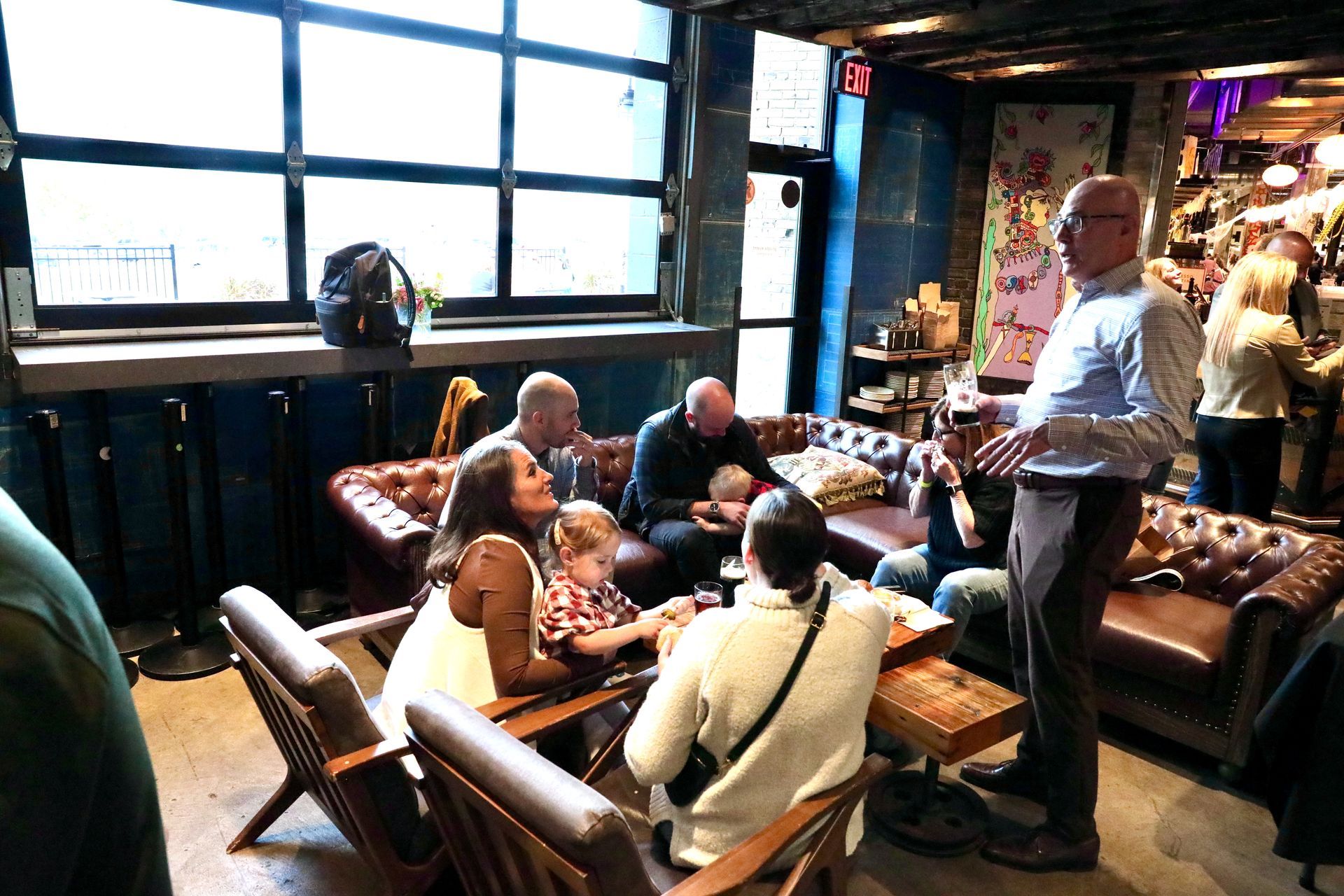 People gathered in a bar: some seated on couches and chairs, others standing and talking.