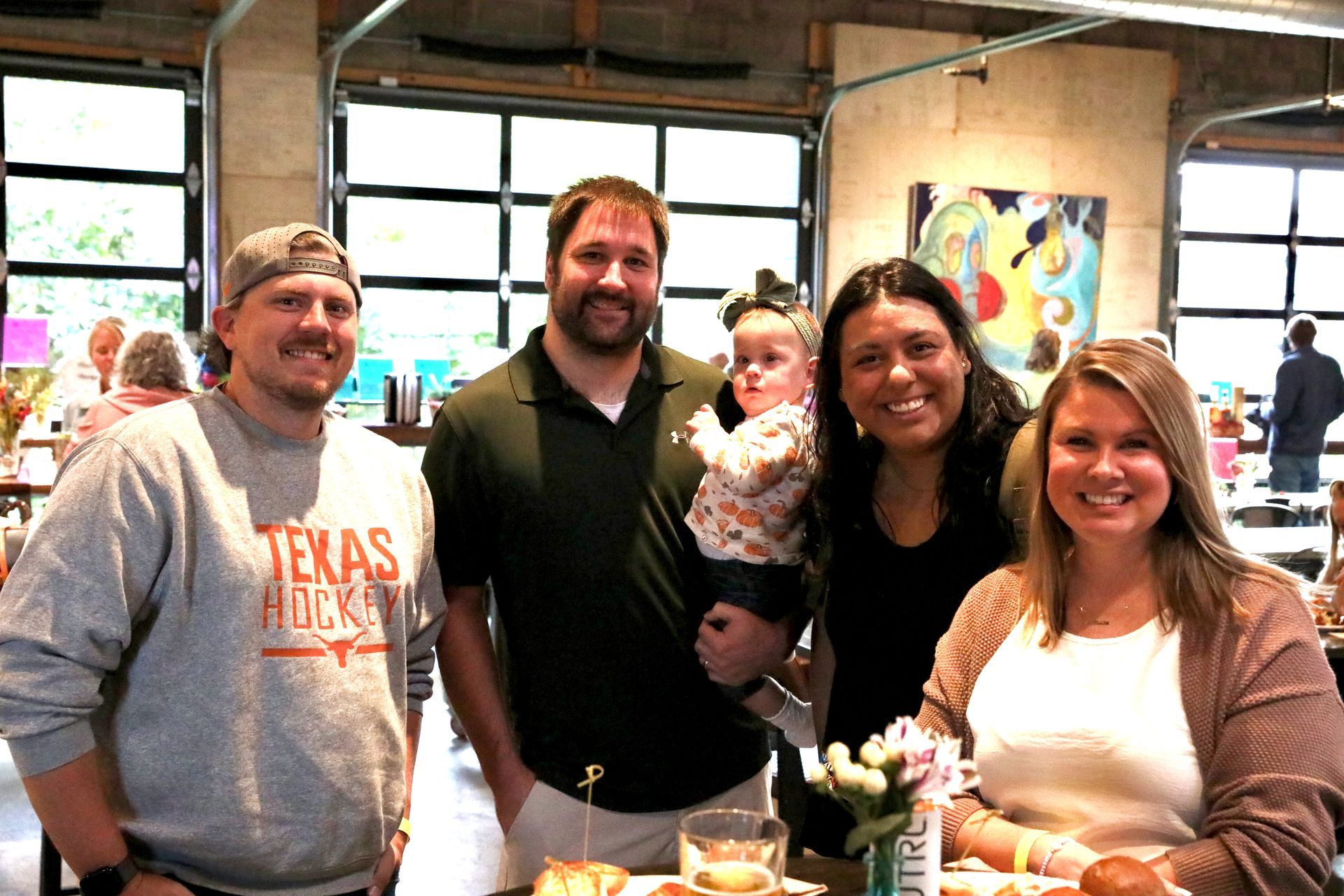 Group of people smiling, posing for photo inside building; tables, artwork, and open garage doors visible.