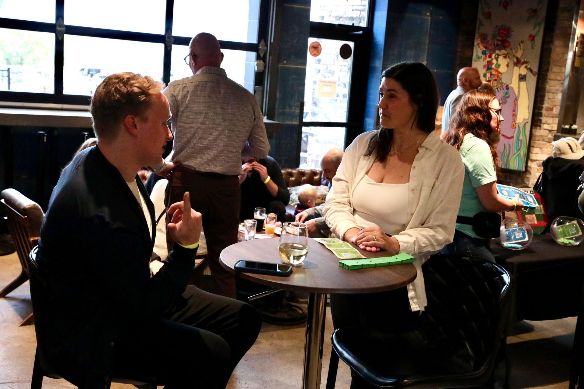 Two people seated at a small table, talking in a dimly lit bar. Several other people in background.