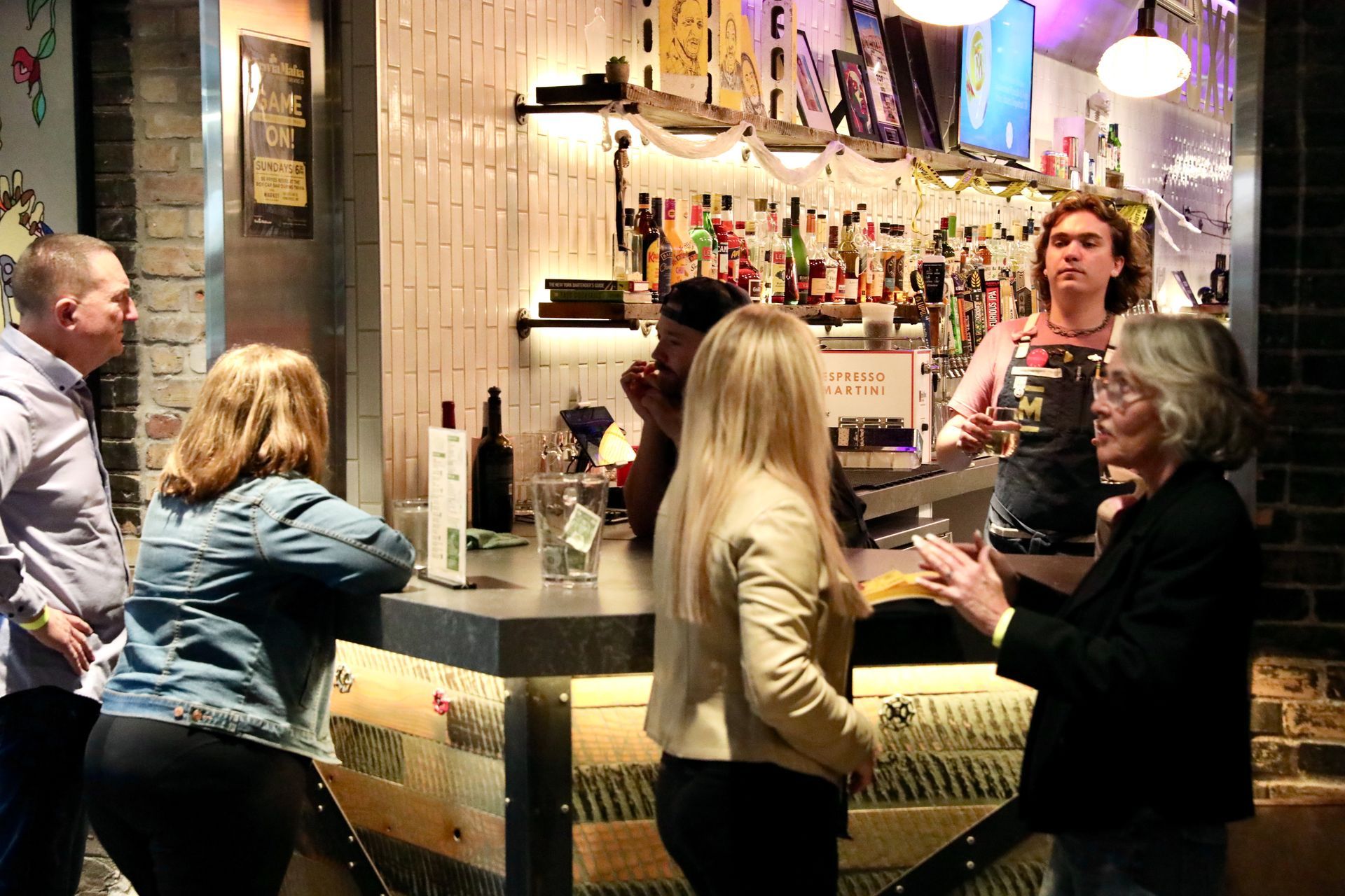 People at a bar, chatting with a bartender. Interior setting with drinks and bar shelves visible.
