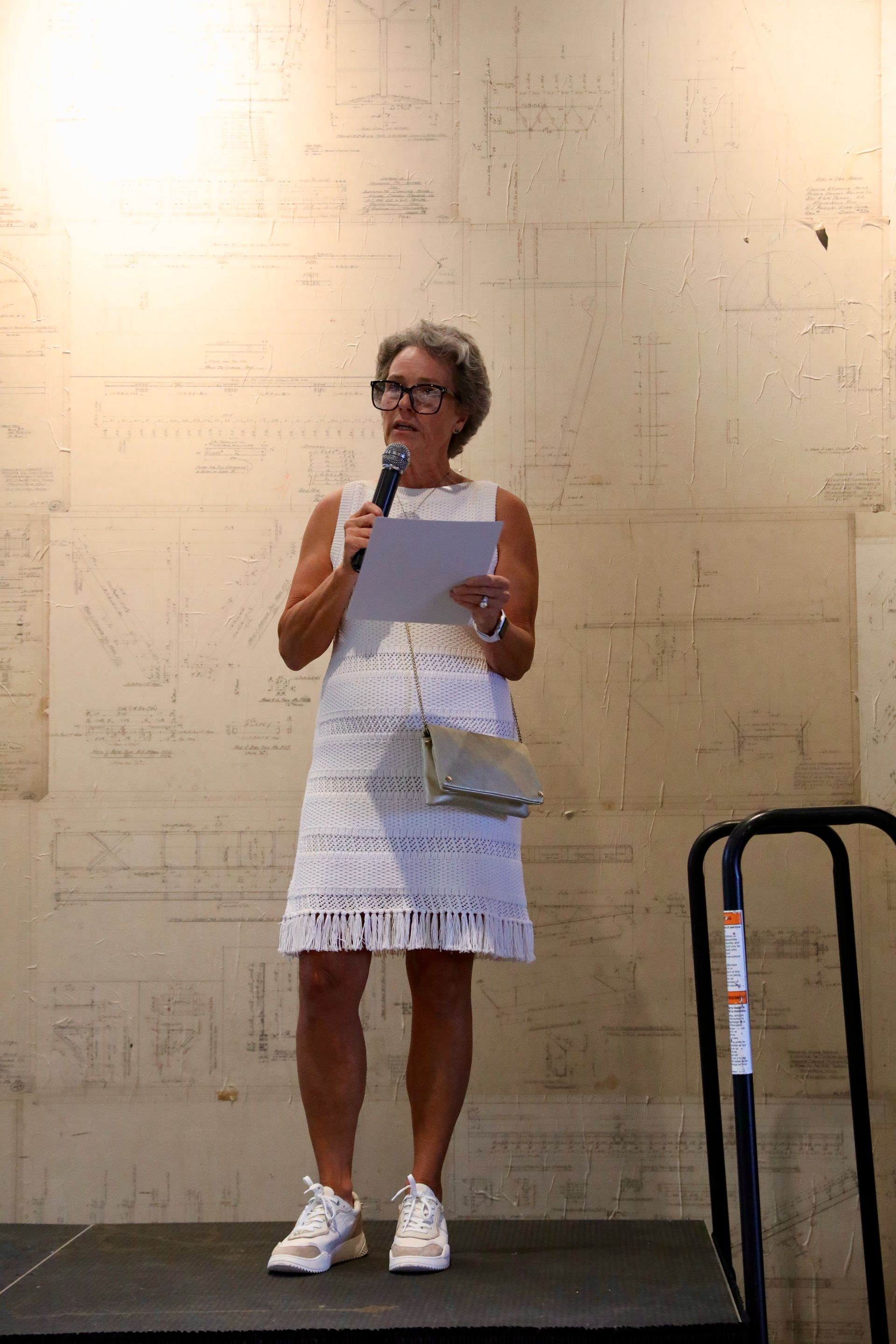 Woman speaking at a podium, holding a microphone and papers. White dress and shoes, gold purse. Wall with drawings in background.