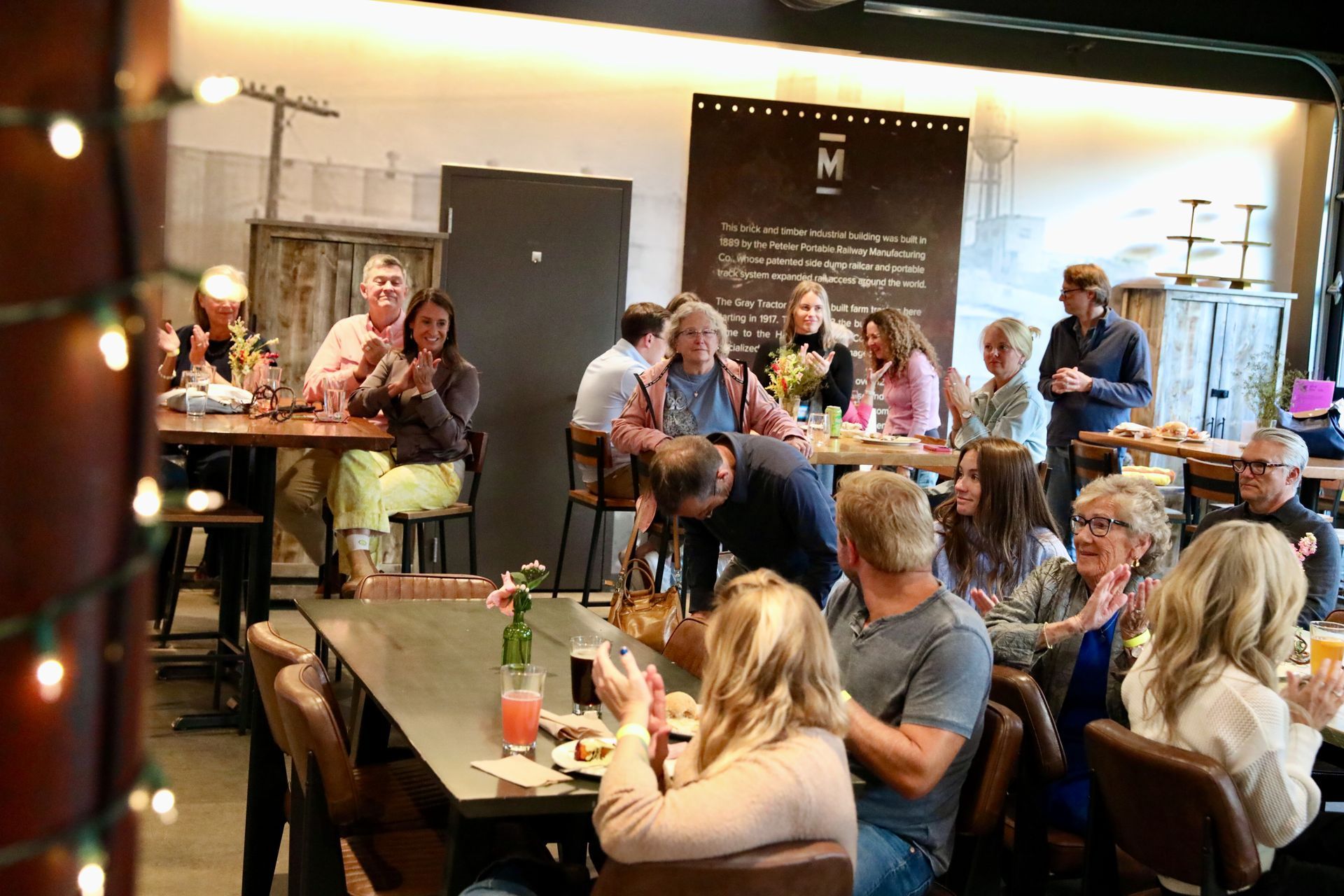 People inside a restaurant clapping. Some are seated at tables with drinks and food. A large sign hangs in the background.