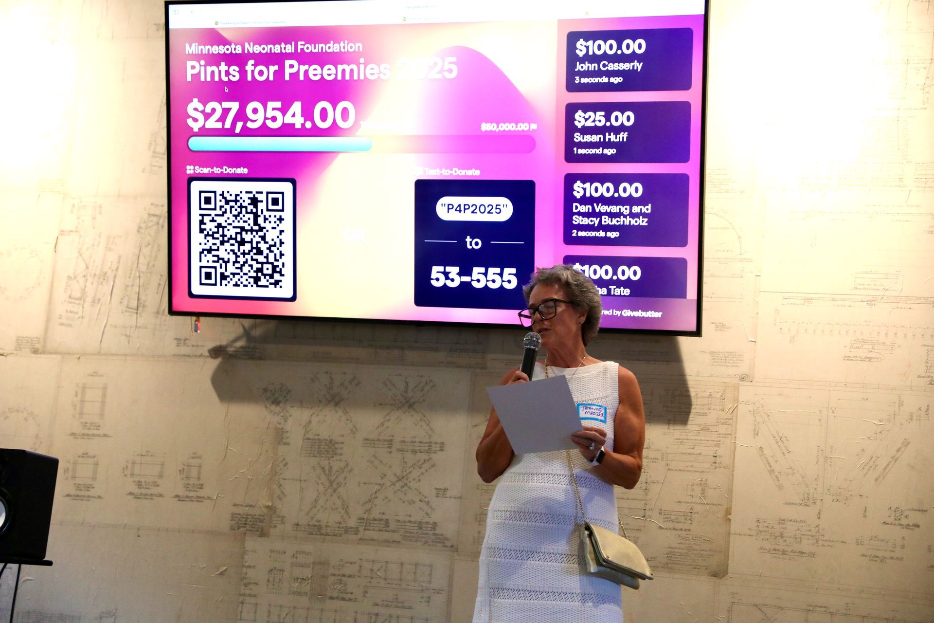 Woman speaking at a fundraiser with a digital display showing donation progress.