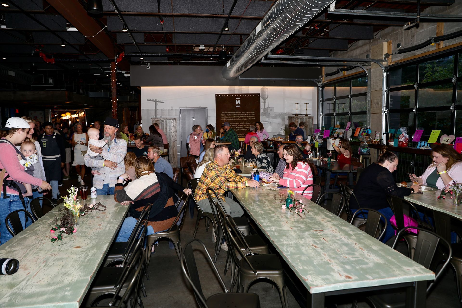 Large gathering at tables inside a venue with many people.  Some are seated, others are standing.  Brightly lit, many smiles.