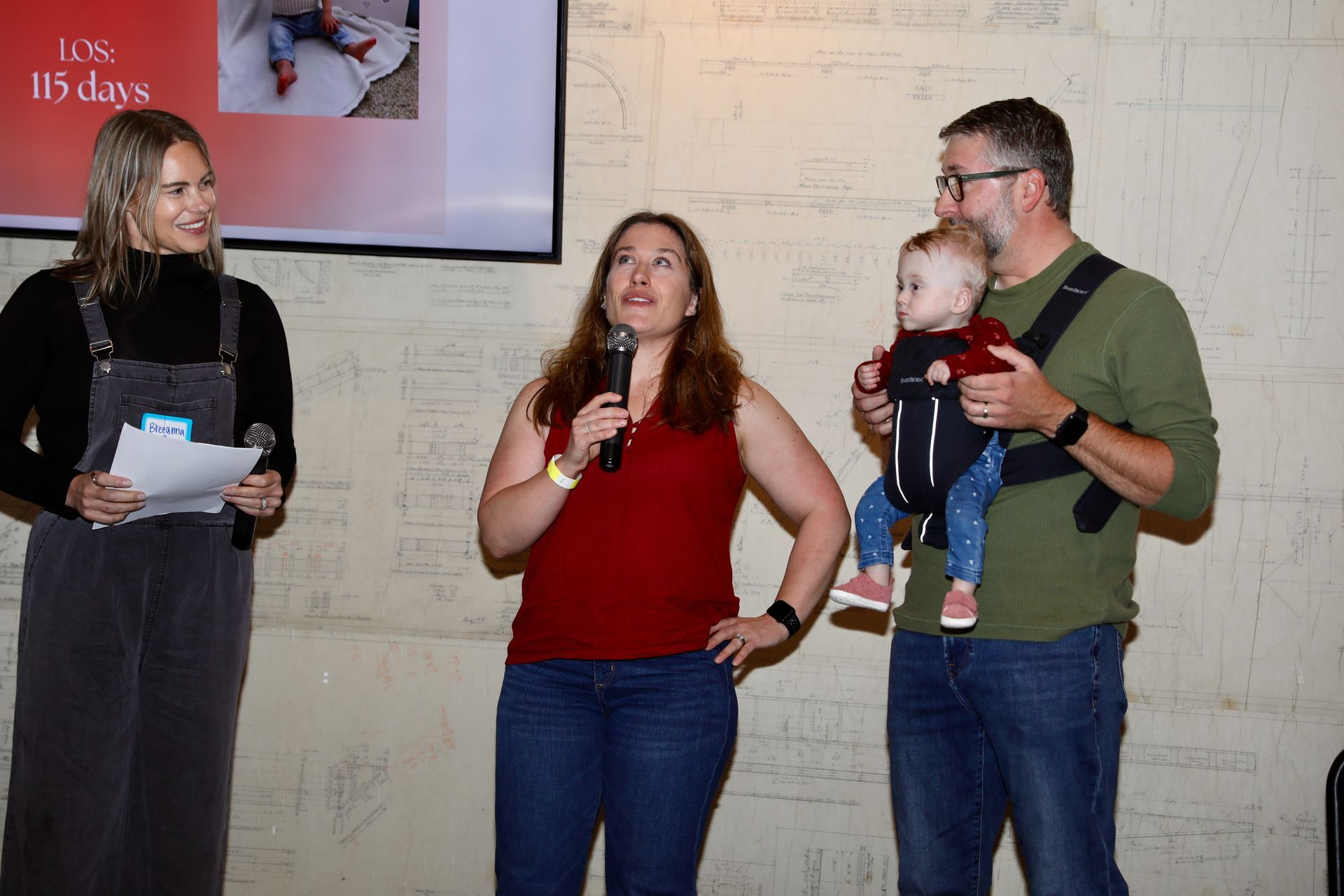 Three people speaking at a presentation. A woman with a microphone, a man holding a baby, and another woman holding papers.
