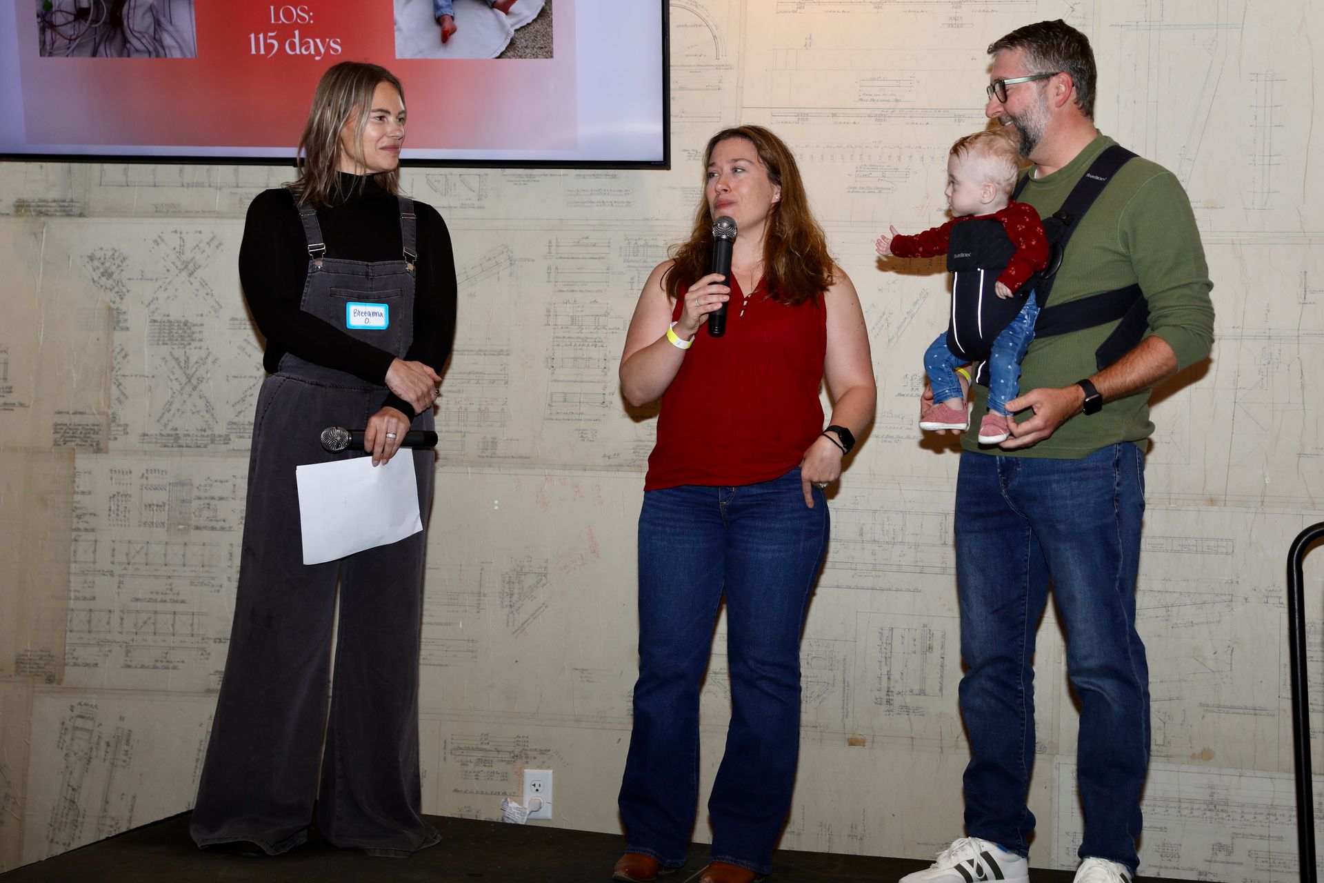 Two women, a man holding a baby, on a stage. One woman speaks into a microphone. They are in front of a wall.
