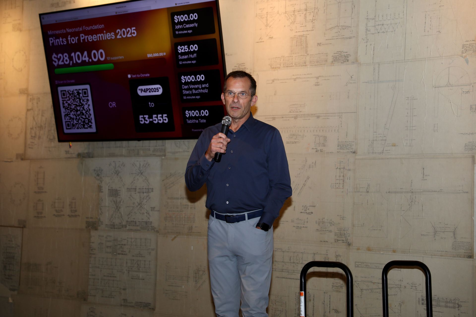Man speaking at an event, stands in front of a screen displaying fundraising progress.
