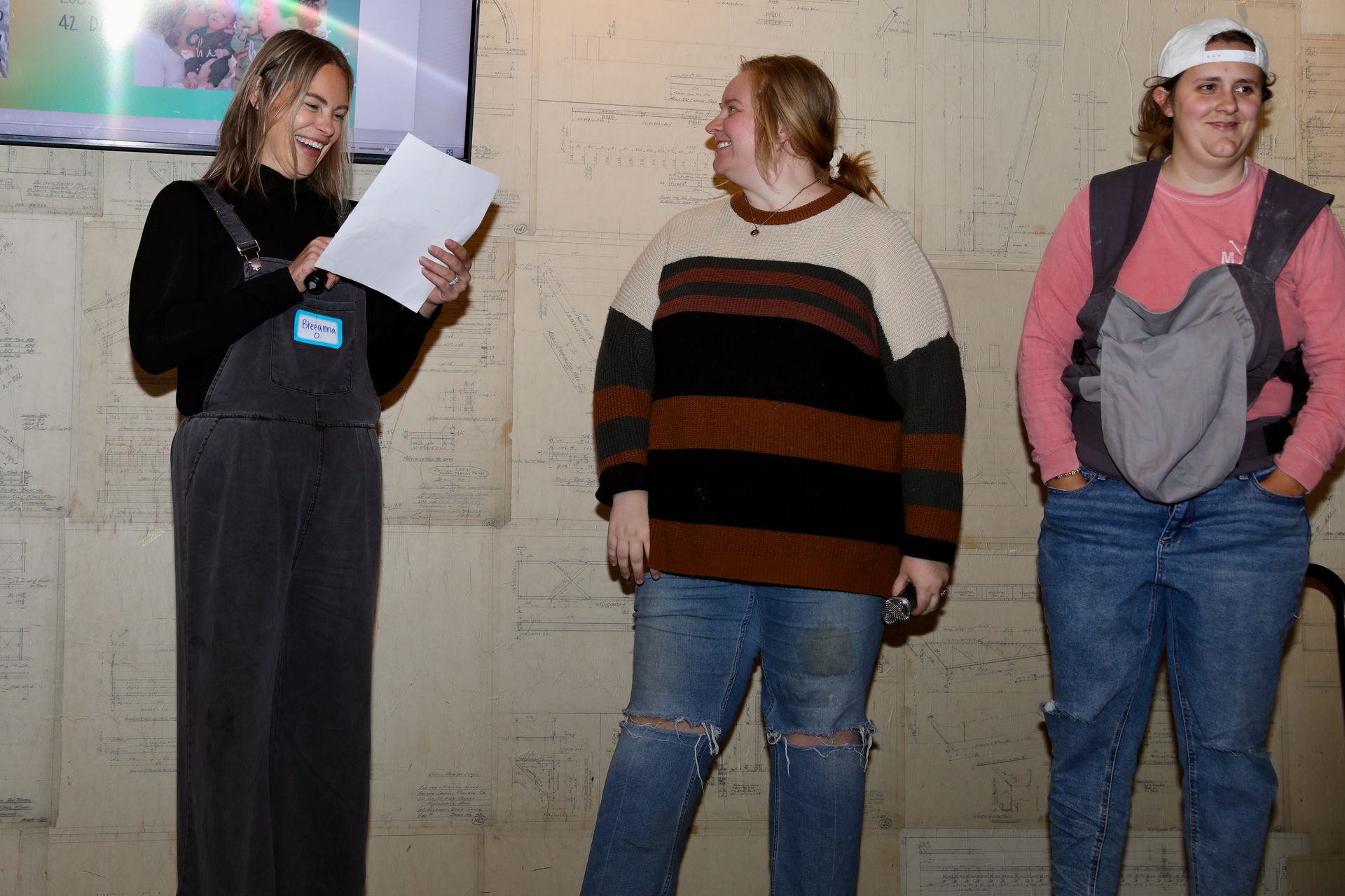 Three people standing in front of a wall with writing. One holds paper and speaks, the others laugh.