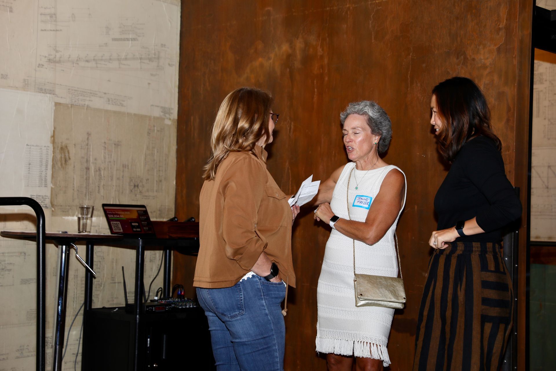 Three people conversing near a stage setup. One wears a white dress, the others casual attire. Wooden backdrop.