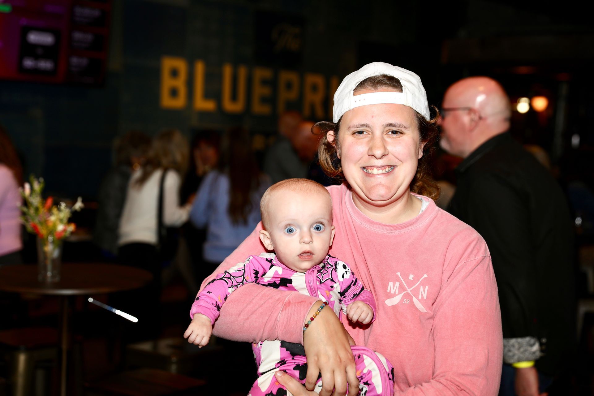Woman in white cap holds a baby wearing pink and white patterned onesie at a bar; 