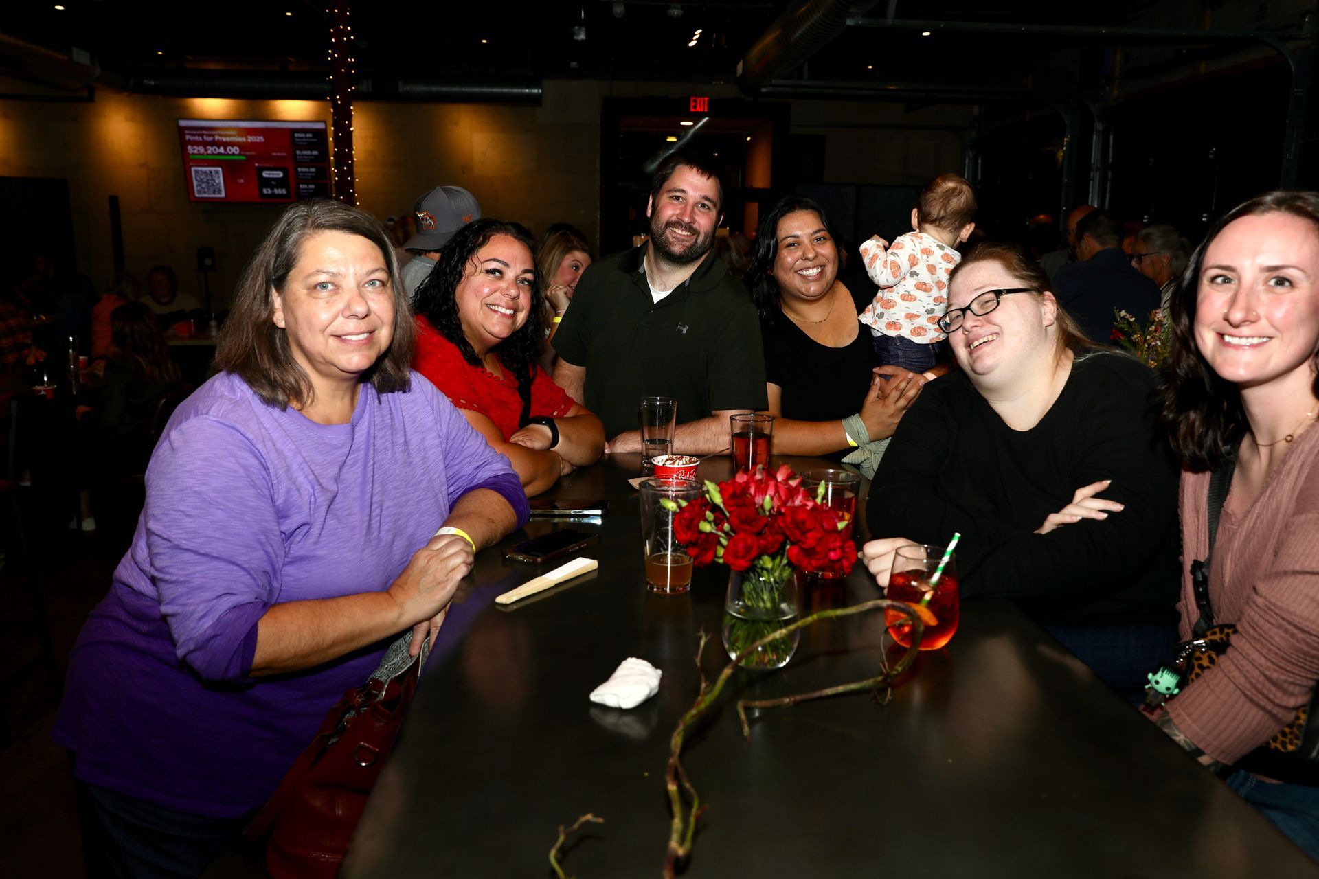 People at a bar, smiling at the camera. Lit bar area with drinks and festive decorations.