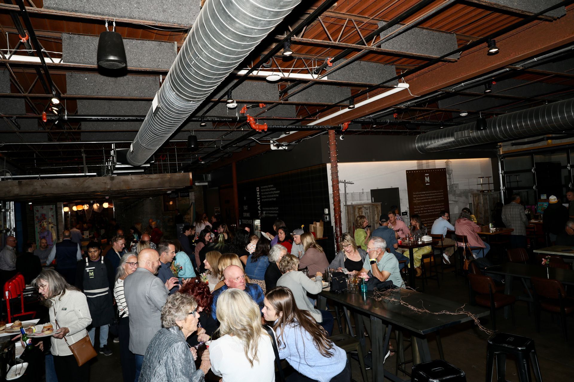 Large crowded event in an industrial-style space with bar and diverse attendees socializing.