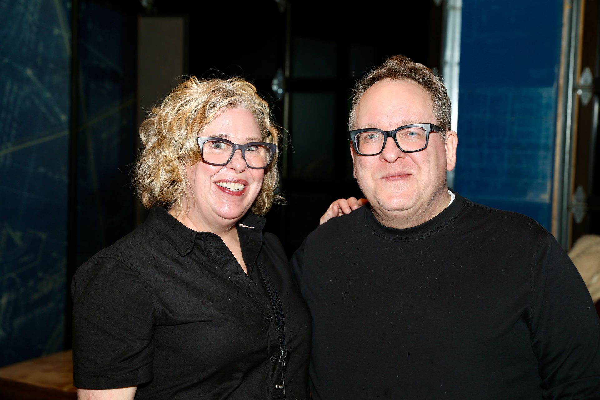 Two people in glasses posing together, smiling. One in a black shirt, the other in a black button-down.