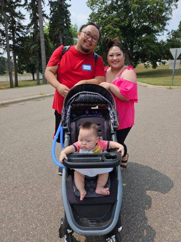 Family of three smiles at camera; baby in stroller. Outdoor setting; asphalt path, trees, and blue sky.