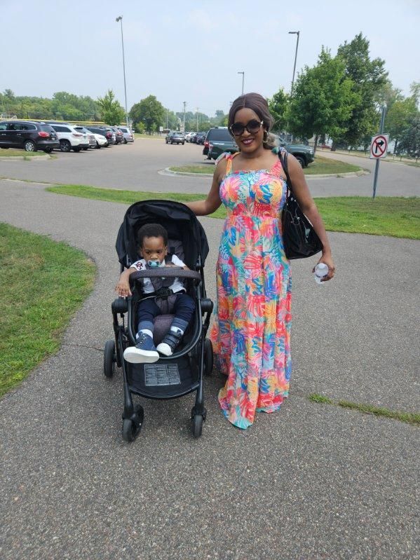 Woman in colorful dress poses with a child in a stroller on a paved path outdoors.