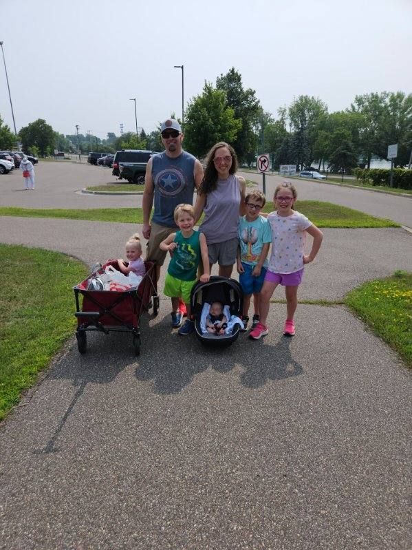 Family of seven poses on a paved path in a parking lot. A child is in a wagon, and another is in a car seat.