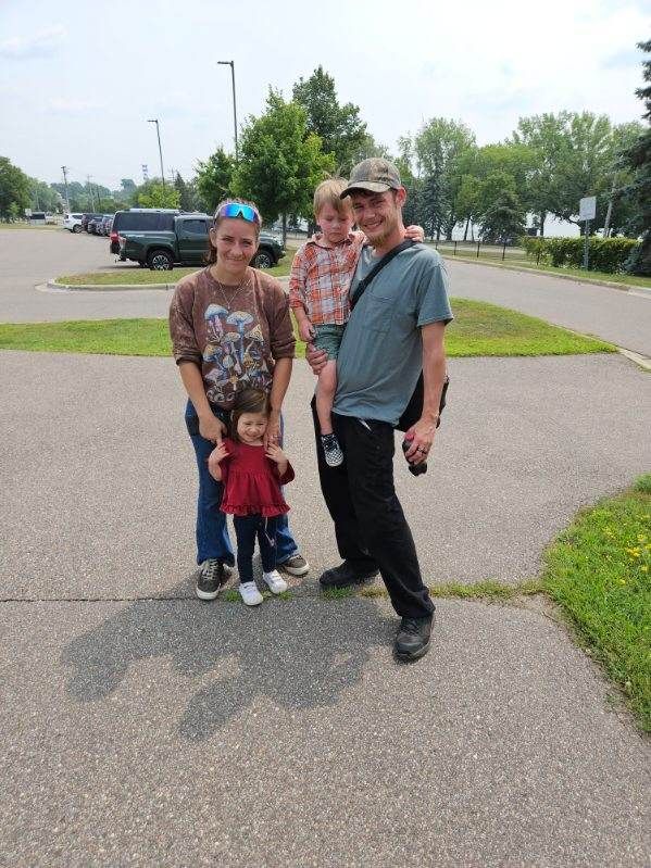 Four people standing outside: a girl in a red dress, two other children and an adult male. Paved area, parked cars.