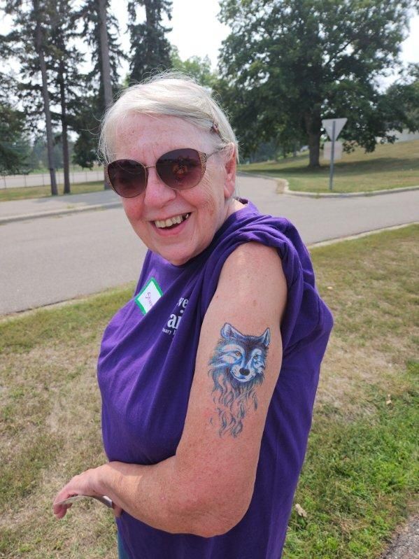 Woman with sunglasses smiles, arm raised to show blue wolf tattoo, wearing purple shirt.